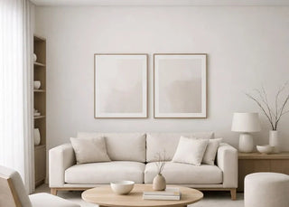 A modern, minimalist living room featuring a light beige sofa, a round wooden coffee table, a matching accent chair, and a large light beige rug on light wood flooring. Two minimalist framed prints hang on the wall above the sofa.